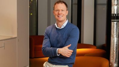 Mark Fairless, smiling, in a blue sweater and beige pants, arms crossed, in a modern office.