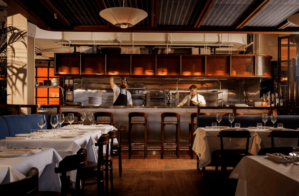 Elegant restaurant interior with chefs working in an open kitchen, set tables, and modern lighting.