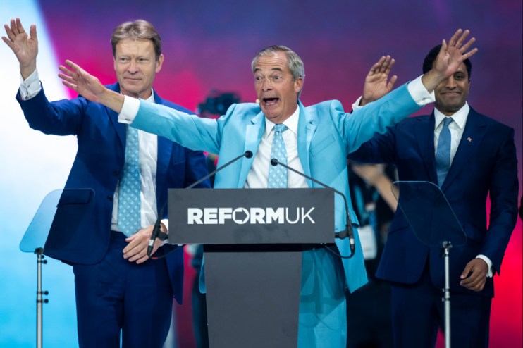 Nigel Farage at a Reform UK podium, arms outstretched, with two men in suits beside him on stage.