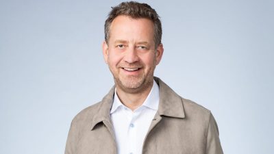 Smiling man with a beard, wearing a light brown jacket, white shirt, and blue jeans, hands in pockets.