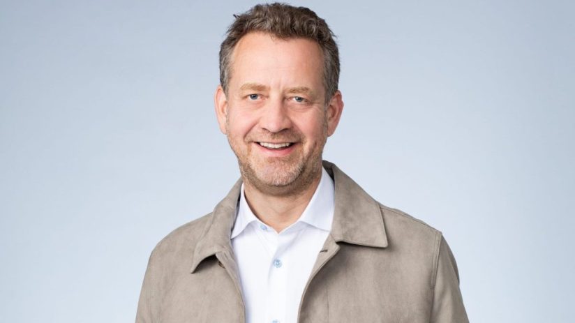 Smiling man with a beard, wearing a light brown jacket, white shirt, and blue jeans, hands in pockets.