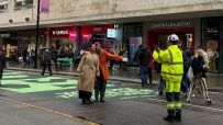 Pedestrians pose on Oxford Streets new green pavement, with a worker in high-vis taking a photo. Mayor of London banner.