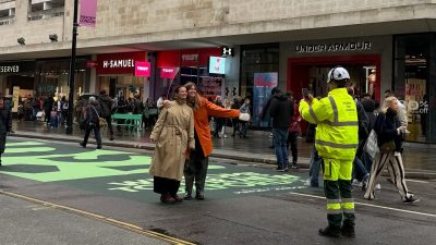 Pedestrians pose on Oxford Streets new green pavement, with a worker in high-vis taking a photo. Mayor of London banner.