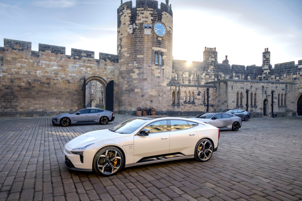White Polestar 5 electric luxury sedan and two grey Polestar 5s parked on cobblestones outside Alnwick Castle, UK.