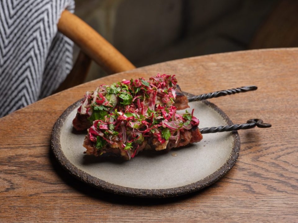 Two Iberico pork souvlaki skewers with pickled red onion and herbs on a grey plate, on a wooden table.