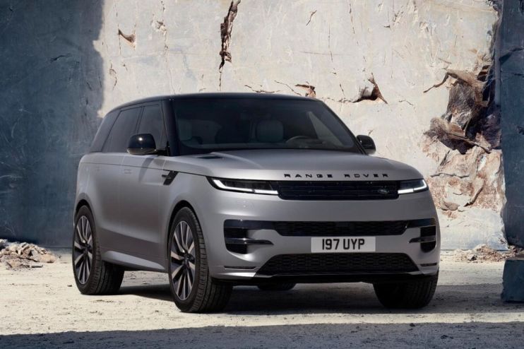 Front view of a silver Range Rover Sport SUV with a black roof, parked against a light-colored rock wall.