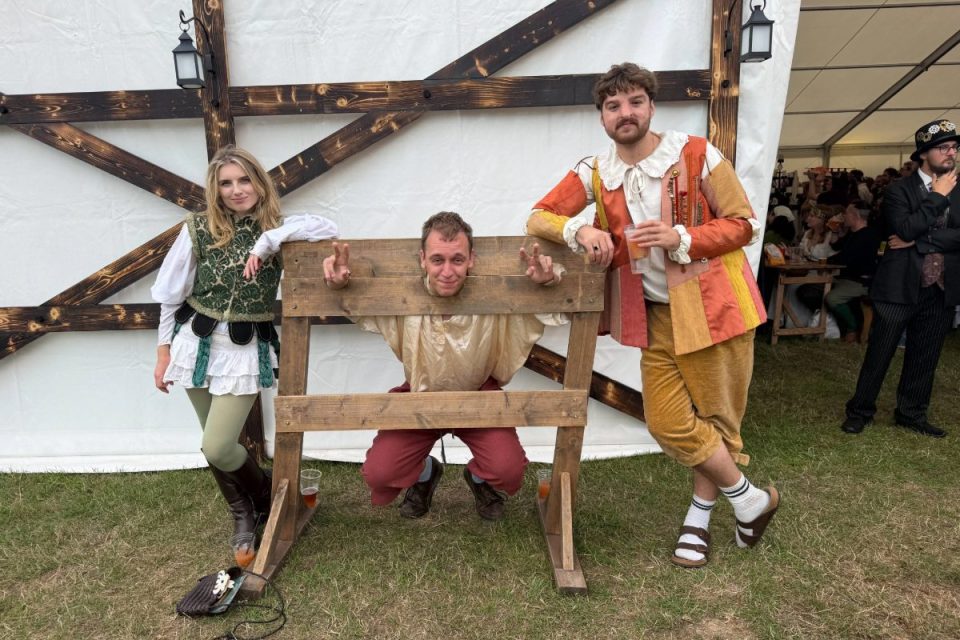 Three attendees in Renaissance costumes posing with a man in stocks at a UK Renaissance Fair.
