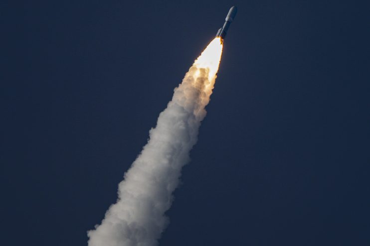 Rocket launch with bright orange exhaust flame and white smoke plume against a dark blue sky