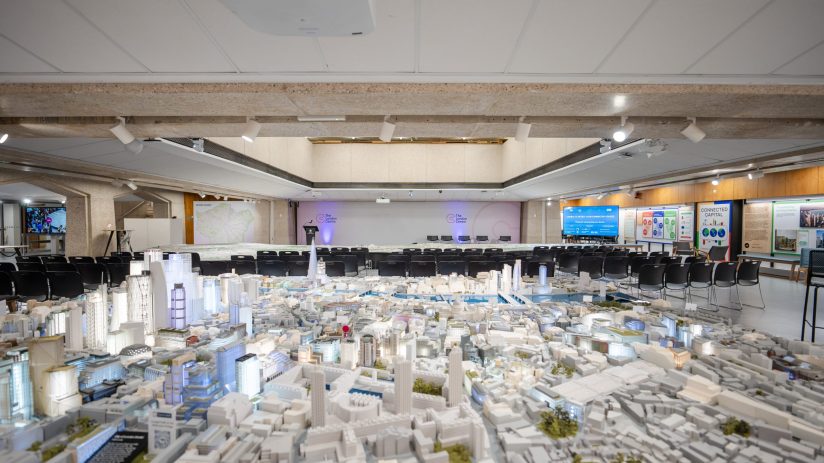 Detailed architectural model of a city, with empty chairs arranged for a presentation in the background.