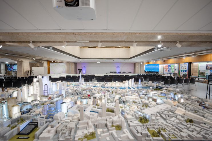 Detailed architectural model of a city, with empty chairs arranged for a presentation in the background.