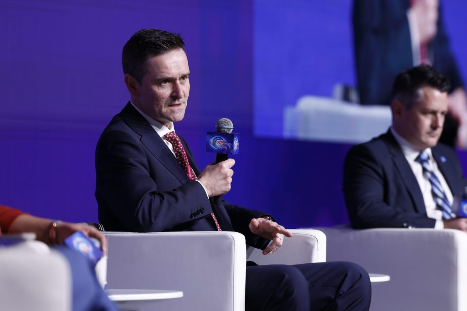 Man in suit speaking into a microphone with GTS Hong Kong logo at a business event.