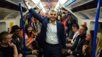 Sadiq Khan smiles, holding a blue grab rail on a crowded London Underground train in 2016, surrounded by diverse commuters.