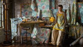 Man in a yellow apron with a floral pattern and tattoos, surrounded by Morris & Co. wallpaper designs.