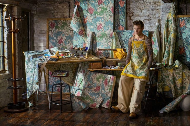 Man in a yellow apron with a floral pattern and tattoos, surrounded by Morris & Co. wallpaper designs.