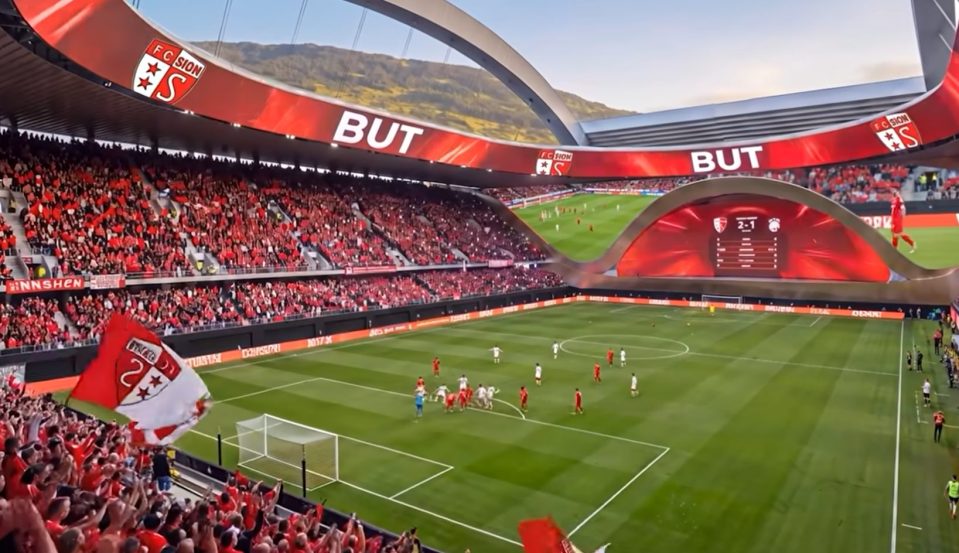 FC Sion football stadium with fans in red, players on the field, and a score of 2-1 on a large screen