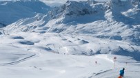 Ski slope with a skier in blue and orange, snow-covered mountains, and a clear sky in the background