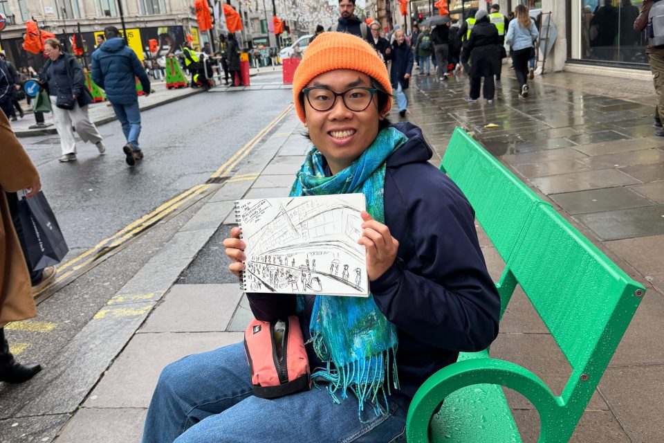Street artist in an orange hat and blue scarf holding a drawing of Oxford Street pedestrians.