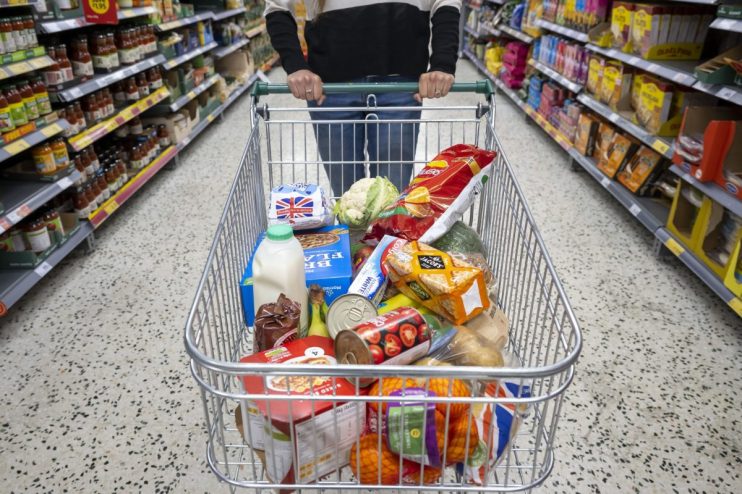 Shopper pushing a supermarket trolley full of groceries, including milk, vegetables, and packaged foods, down an aisle.