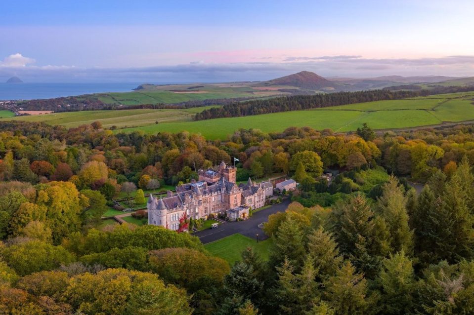 Aerial view of a grand Scottish castle surrounded by autumn trees, green fields, and coastline.
