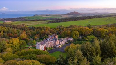 Aerial view of a historic Scottish castle surrounded by autumn trees, with coastline and hills in the Lowlands background.