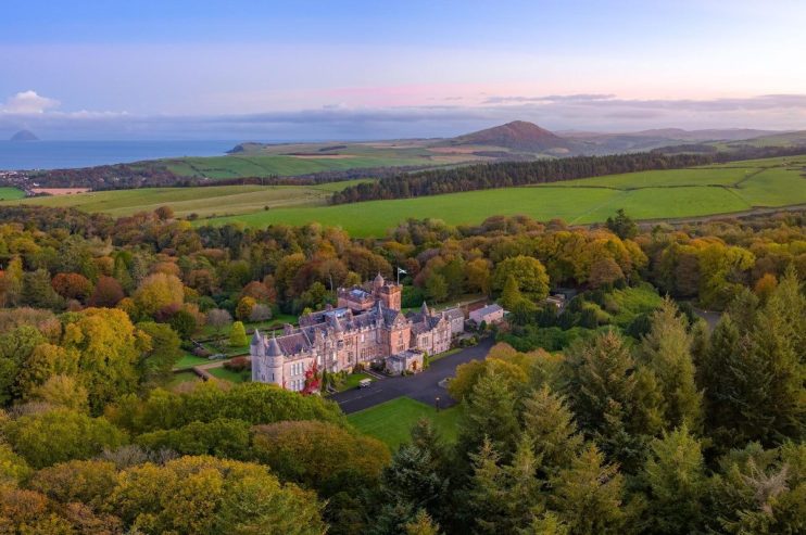 Aerial view of a historic Scottish castle surrounded by autumn trees, with coastline and hills in the Lowlands background.