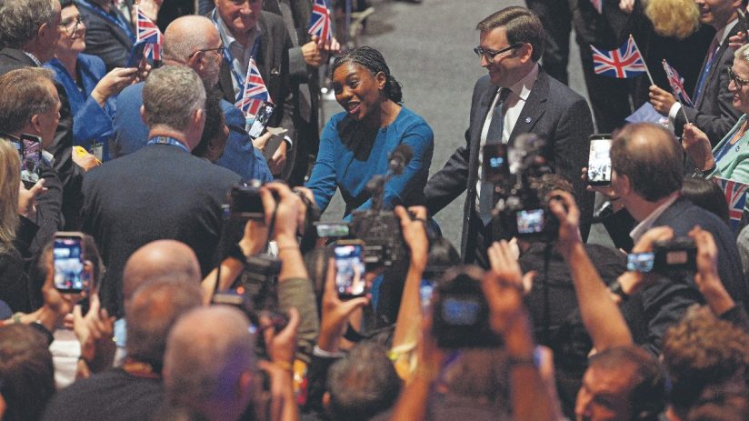 Kemi Badenoch in blue dress at Tory Conference, surrounded by smiling attendees and media filming with phones.