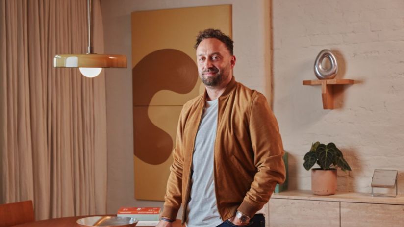 Man in a brown suede jacket standing in a modern room with abstract art, a unique pendant light, and minimalist decor.