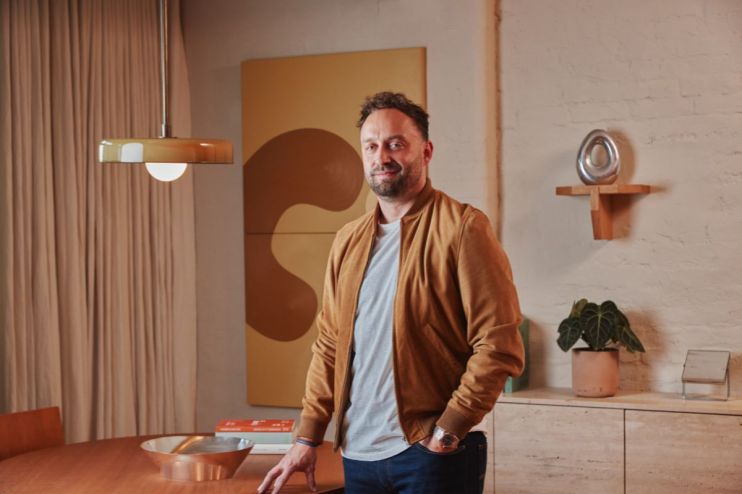 Man in a brown suede jacket standing in a modern room with abstract art, a unique pendant light, and minimalist decor.