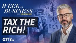 Christian May in glasses with Tax the Rich text, protesting crowd, and signs in background for business news.