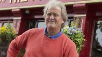 Wetherspoon chairman Tim Martin smiling in a red sweater outside a pub with hanging baskets