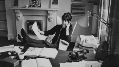 Woman in black dress talking on a rotary phone, feet up on desk, working from home office.