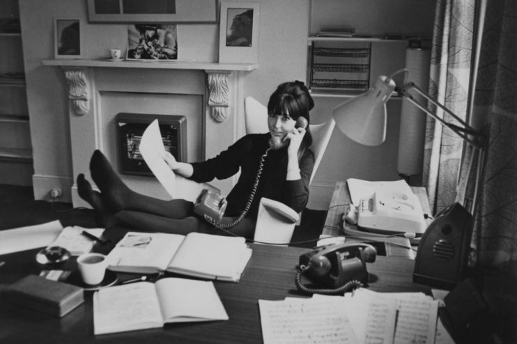 Woman in black dress talking on a rotary phone, feet up on desk, working from home office.