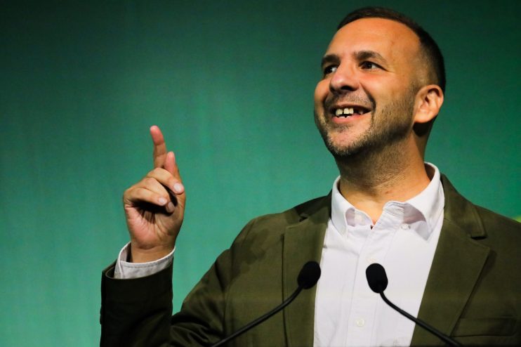 Zack Polanski speaking at a podium, smiling and pointing upwards with his right hand, against a green background.