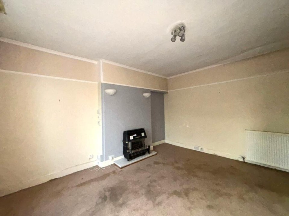 Empty, dated living room in Aberdeen with beige walls, gas fireplace, wall lights, and stained brown carpet.
