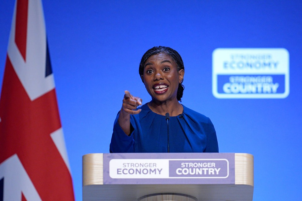 Kemi Badenoch speaks at a podium, pointing, with a Union Jack flag and Stronger Economy, Stronger Country backdrop