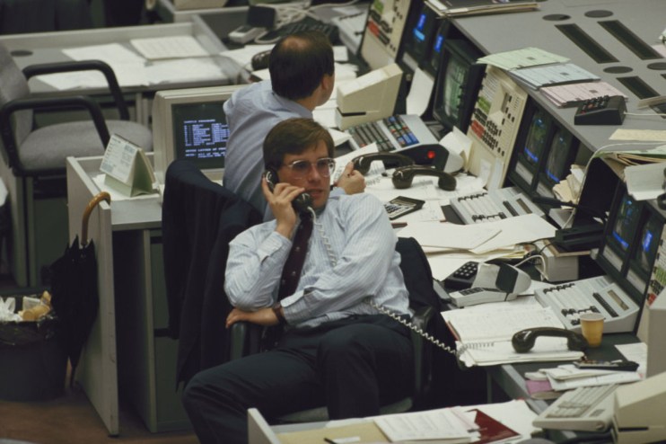 A trader in a striped shirt and glasses on the phone at a busy desk in the City of London during the Big Bang.