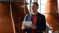 Man in a suit holding a Port Ellen Established 1825 brochure, standing inside a distillery with copper stills.