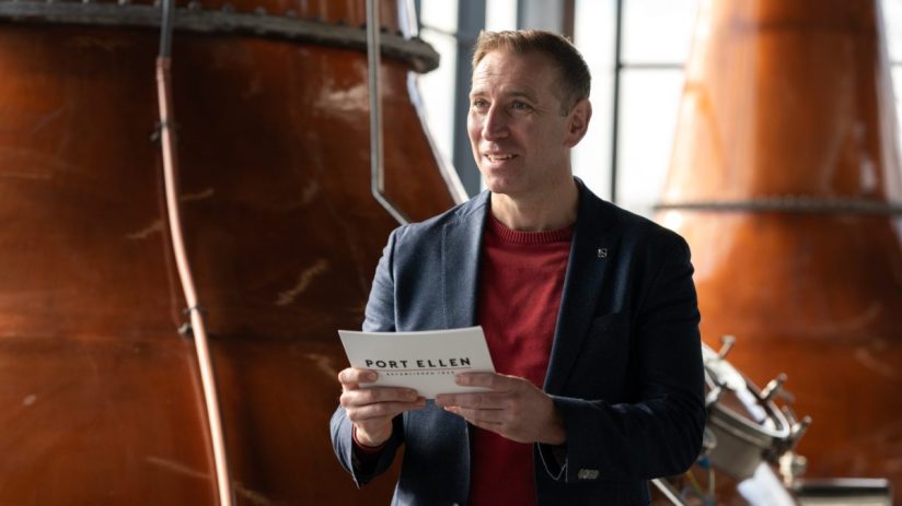 Man in a suit holding a Port Ellen Established 1825 brochure, standing inside a distillery with copper stills.