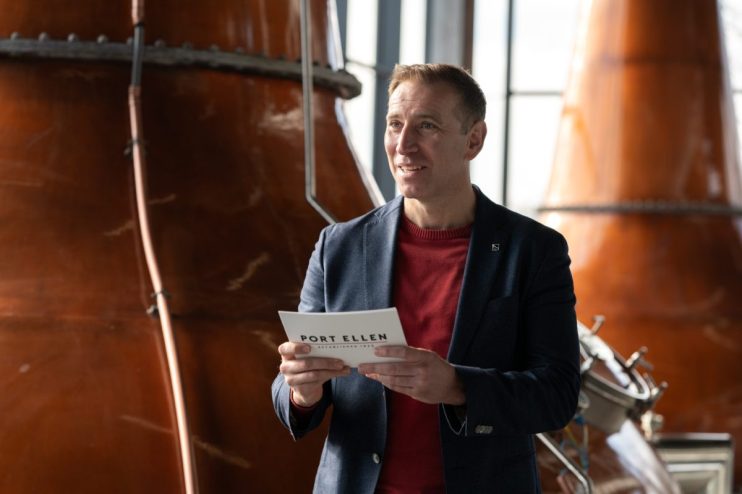 Man in a suit holding a Port Ellen Established 1825 brochure, standing inside a distillery with copper stills.