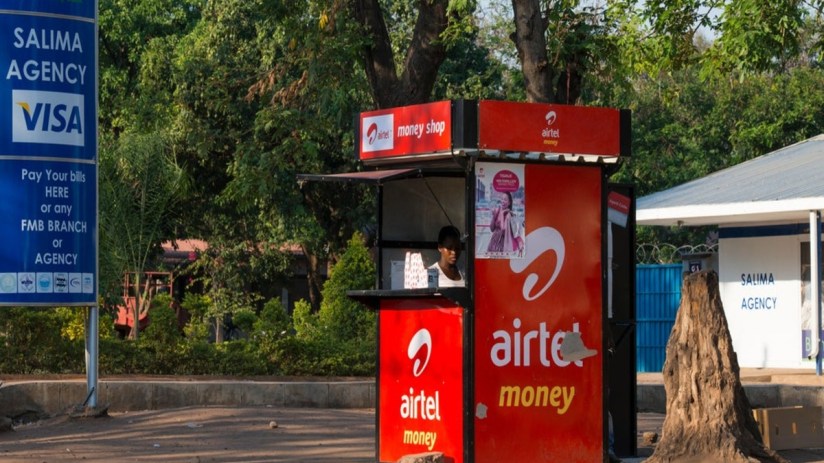 Airtel Money kiosk with a vendor, FMB Salima Agency sign, and lush green trees in the background.