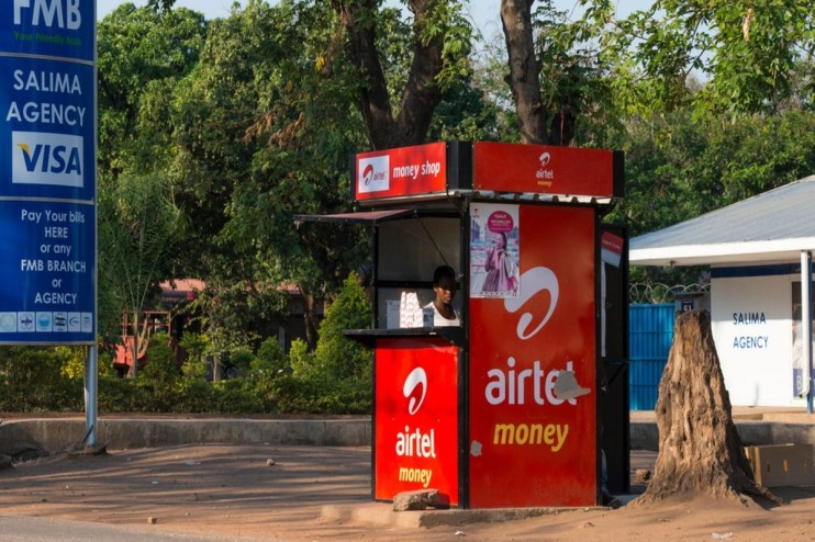 Airtel Money kiosk with a vendor, FMB Salima Agency sign, and lush green trees in the background.