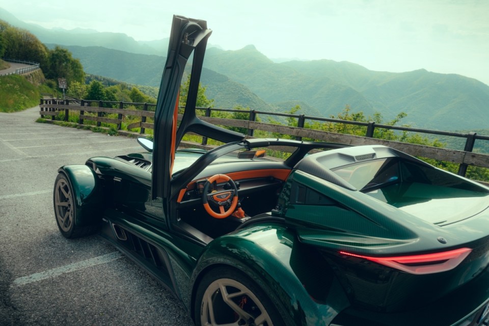 Green Donkervoort sports car with an open door, orange interior, parked on a mountain road with scenic views.