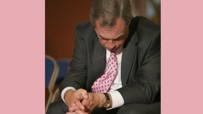 Nigel Farage in a dark suit and pink polka dot tie, looking down with clasped hands, wearing a gold watch