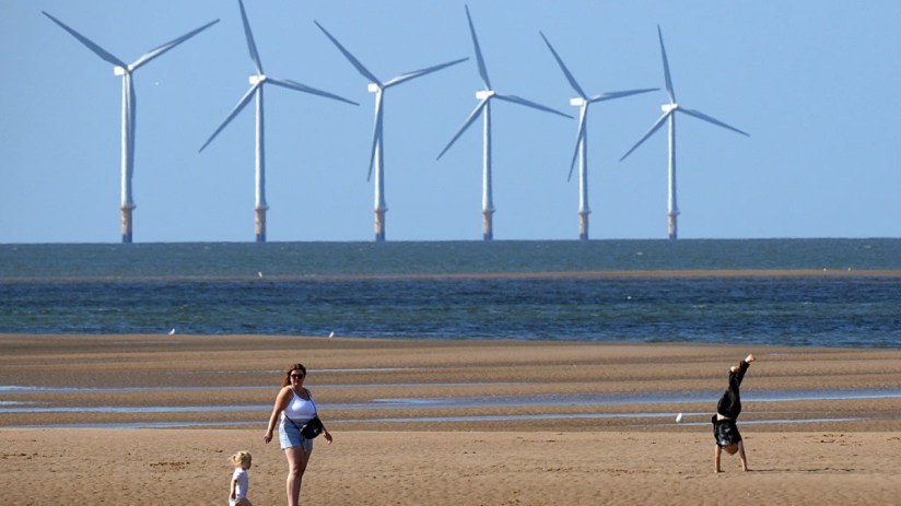 Offshore wind farm turbines visible from a sandy beach with people enjoying the shore.