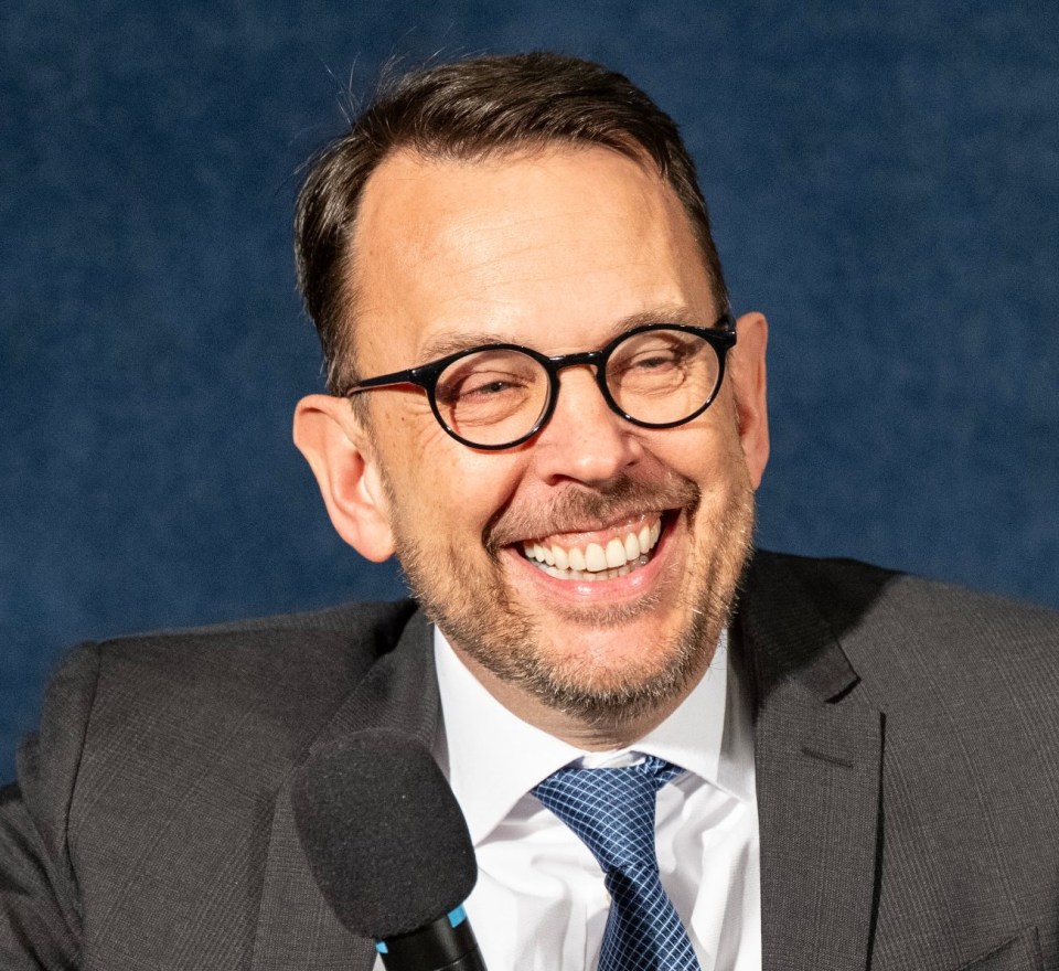 A smiling man in glasses, suit, and tie, speaking into a microphone about Windows Edge browser issues.