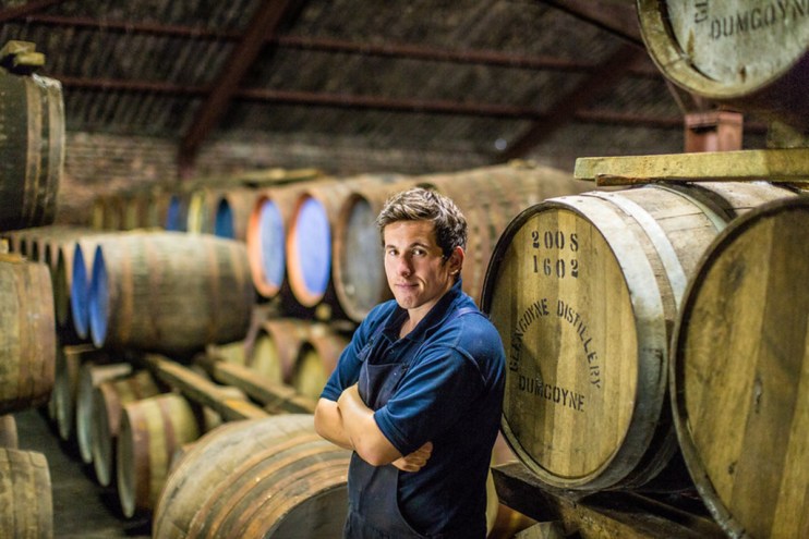 Man in a blue shirt and apron standing among whisky barrels at Glengoyne Distillery, Dumgoyne.