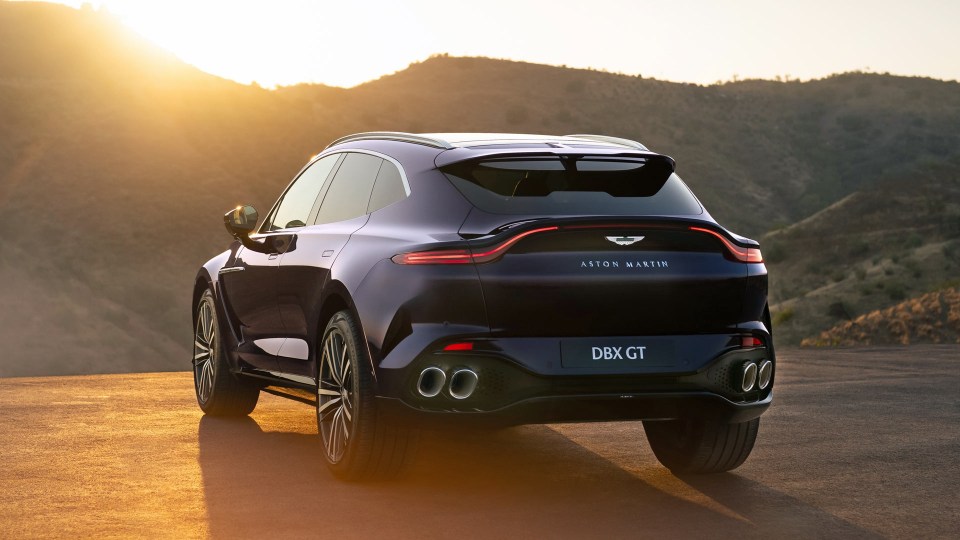 Dark blue Aston Martin DBX GT SUV at sunset on a mountain road, rear view showing taillights and exhaust.