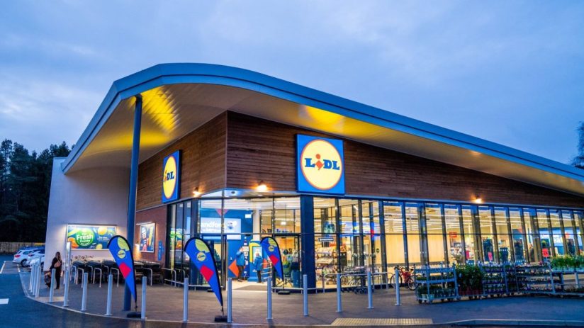 Lidl supermarket exterior at dusk with prominent logo, modern architecture, and customer walking towards shopping carts.