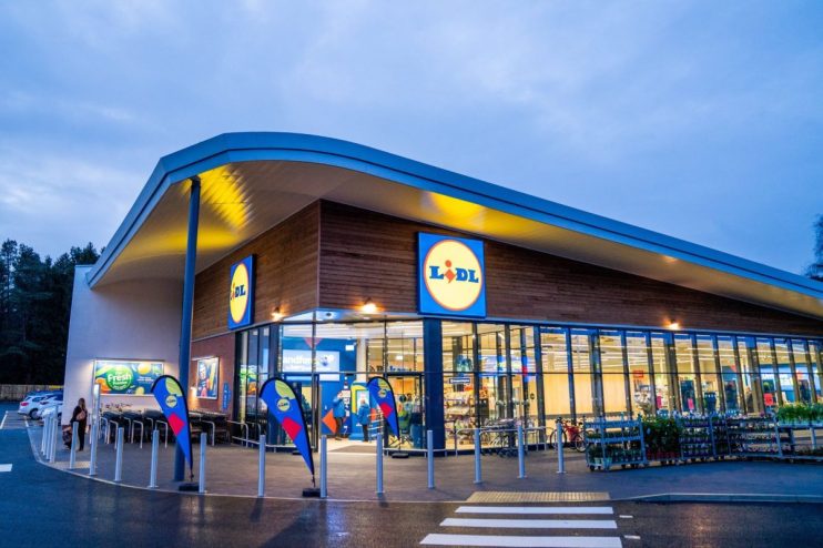 Lidl supermarket exterior at dusk with prominent logo, modern architecture, and customer walking towards shopping carts.