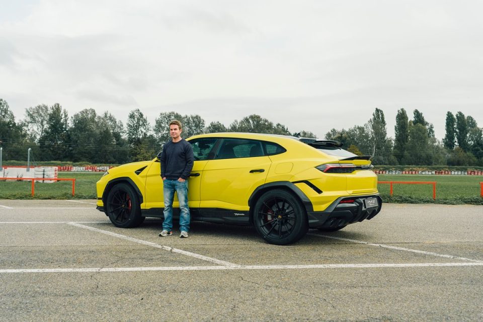 A man in a dark sweater and jeans stands beside a bright yellow luxury SUV on an outdoor track.